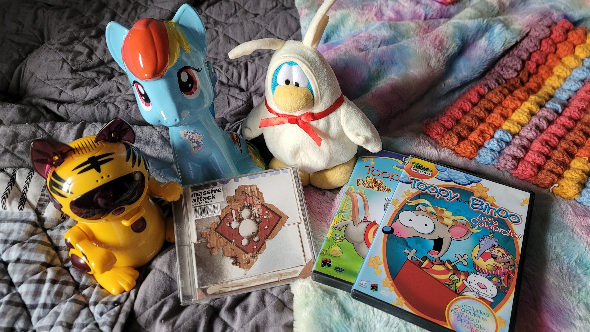 Picture showing a Tiger Electronics orange striped Meow-chi, a My Little Pony Rainbow Dash piggy bank, a CD of Massive Attack's album 'Protection,' a Club Penguin plush of a blue penguin dressed as a white chocolate bunny, and two Toopy and Binoo DVDs