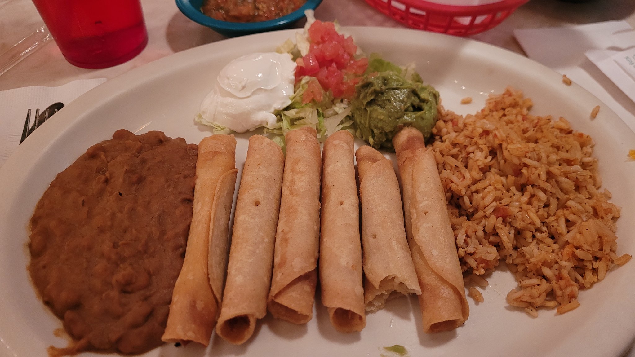 A plate of tex mex food, including chicken flautas, refried beans, Mexican rice, and salad, with sour cream and guacamole on the side